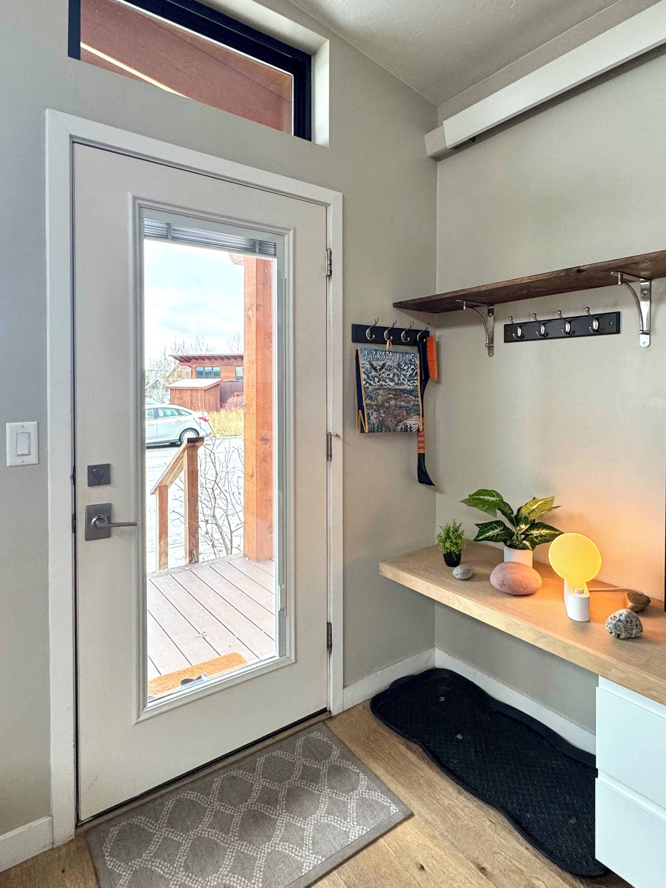 Una entrada moderna con puerta de vidrio, terraza de madera, estante con plantas y perchero en la pared.