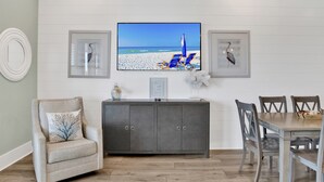 Una sala de estar con un televisor montado en la pared, un mueble gris, un sillón color beige y una mesa de comedor con cuatro sillas.