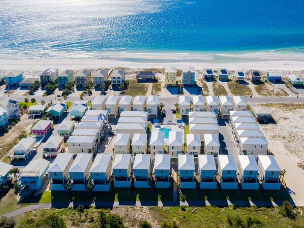 An aerial view of a coastal resort with multiple bungalow-style buildings arranged in rows, a central swimming pool, and a beach in the background.