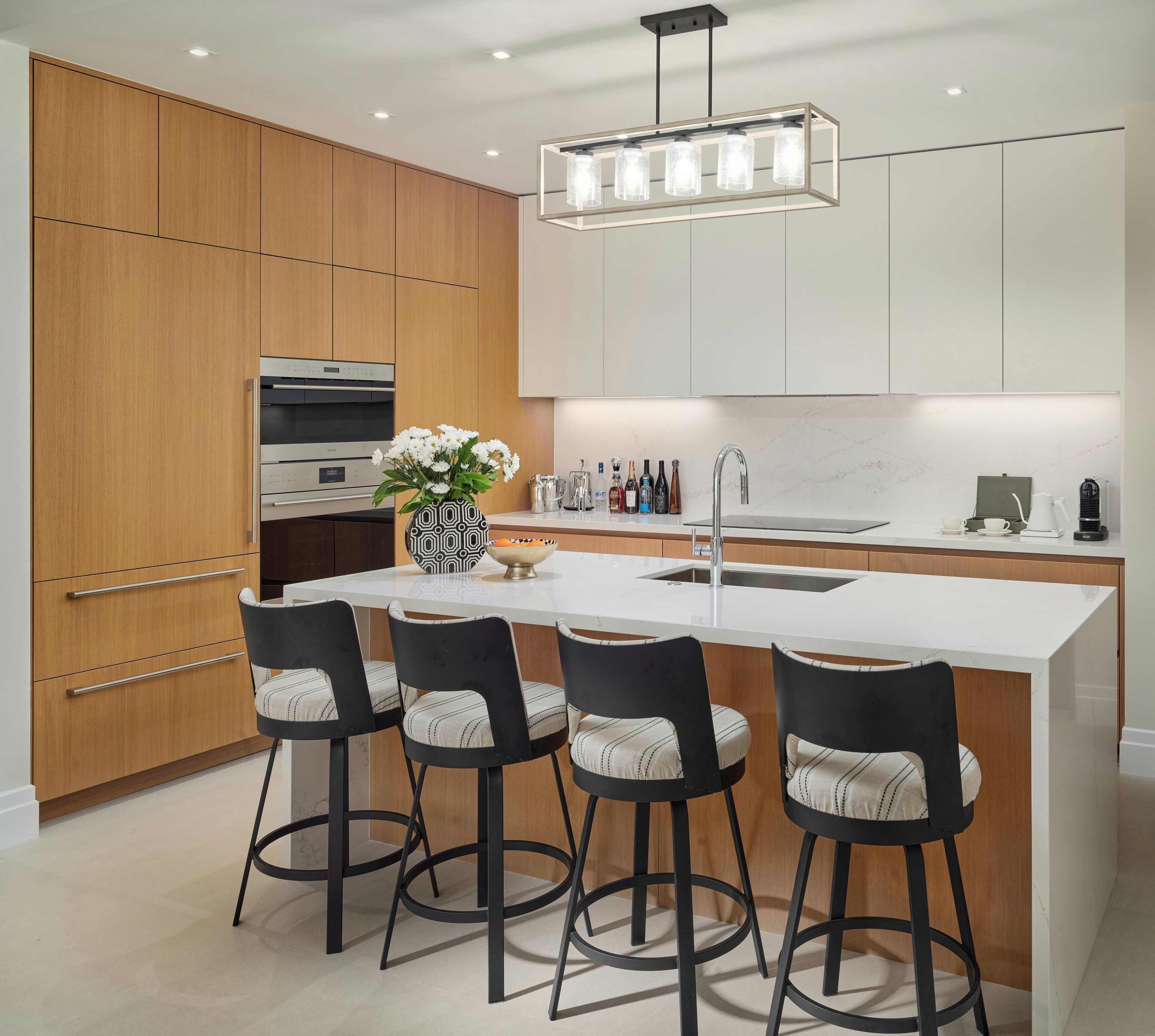 A modern kitchen with wooden cabinets, a central island with bar stools, a built-in oven, and a pendant light fixture.