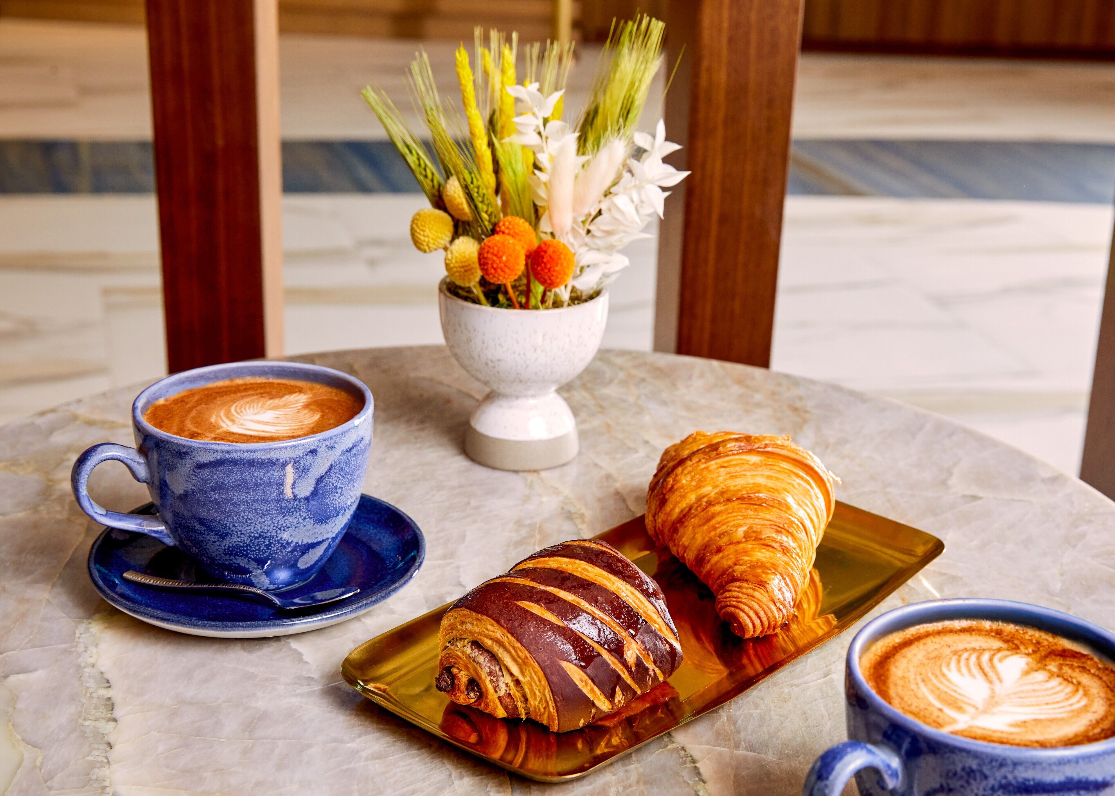 A table with a blue cup of coffee, a croissant, and a pastry on a golden tray, with a white vase of flowers in the background.