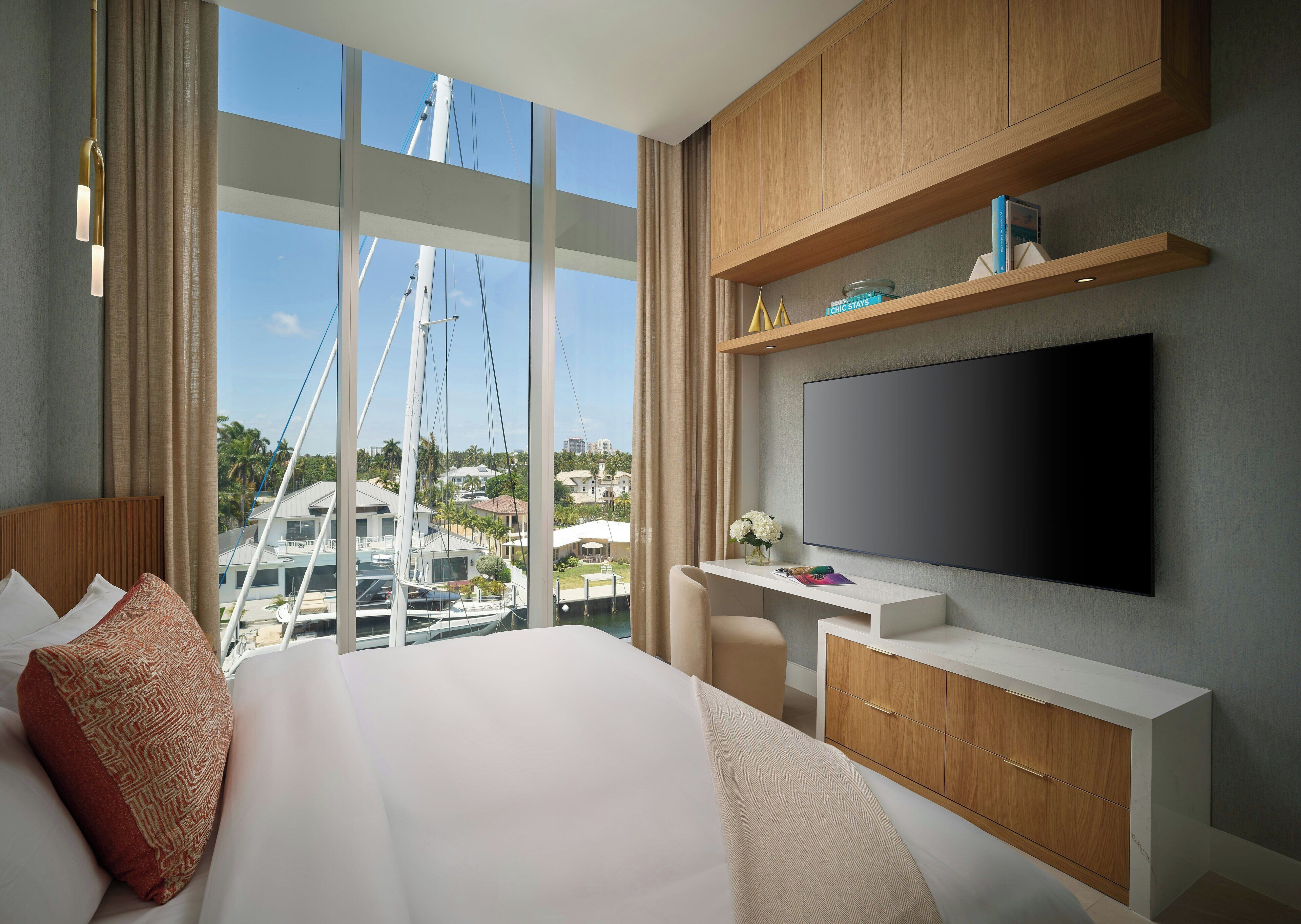 A modern hotel room with a large bed, a desk with a flat-screen TV, wooden cabinets, and a view of boats and buildings.