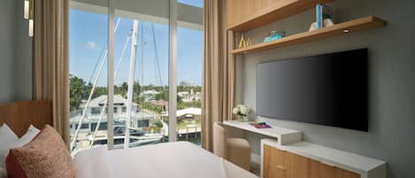 A modern hotel room with a large bed, a desk with a flat-screen TV, wooden cabinets, and a view of boats and buildings.