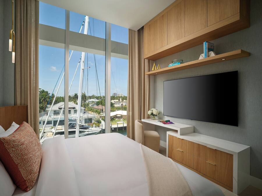 Una moderna camera d'albergo con un letto grande, una scrivania con TV a schermo piatto, armadietti in legno e vista su barche ed edifici.