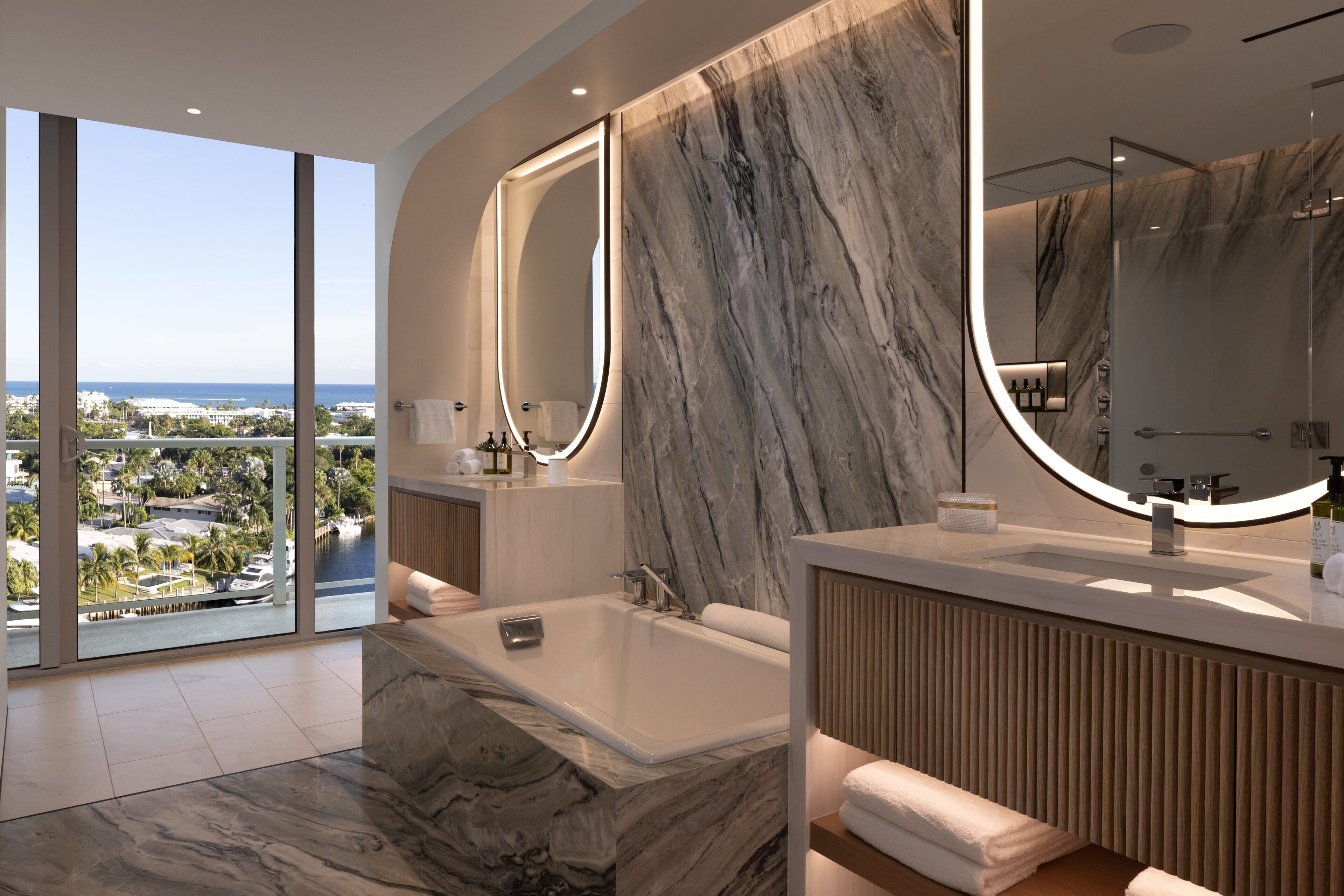 A modern bathroom with a large bathtub, marble walls, and a view of the city.