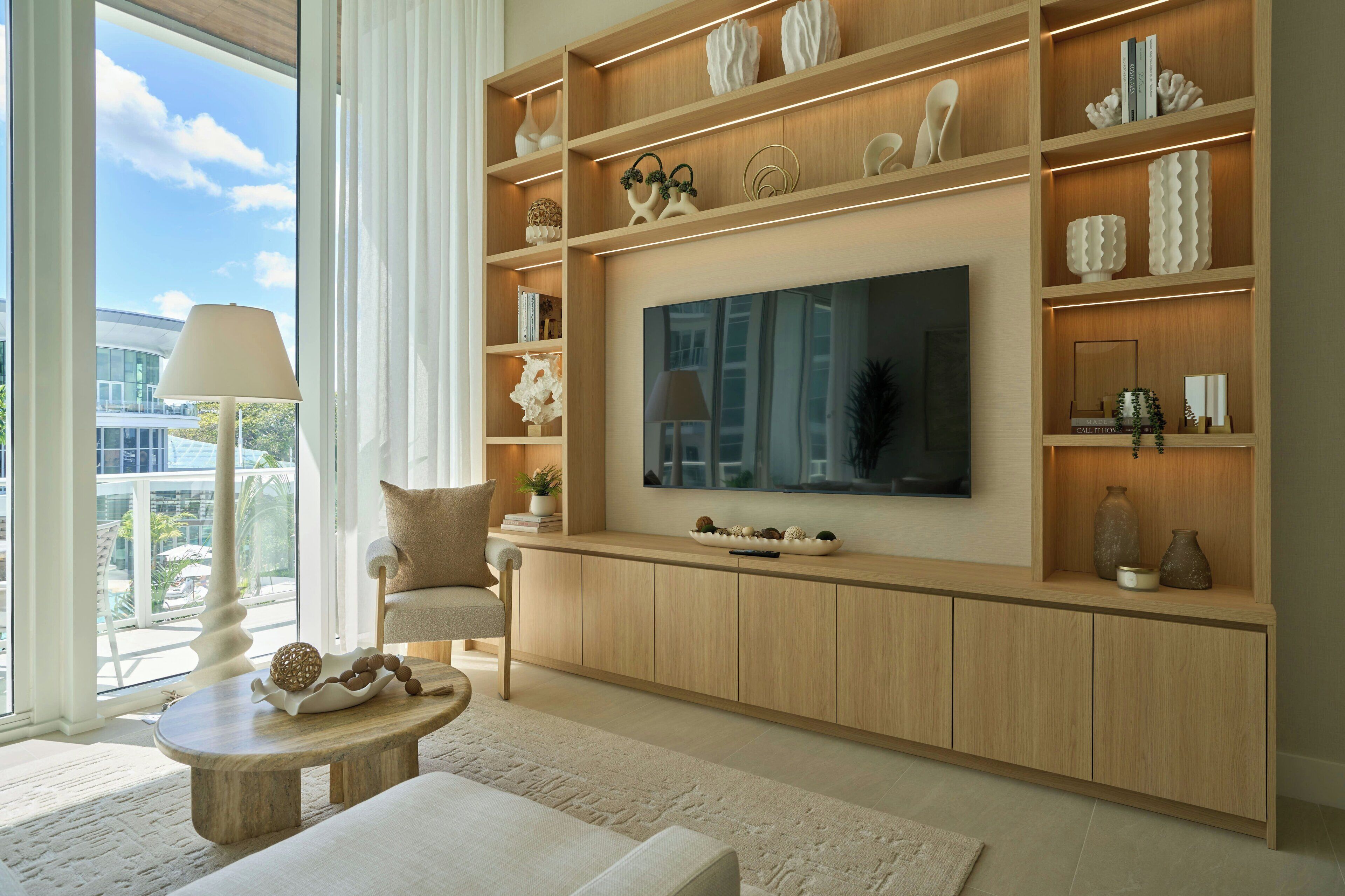 A modern living room with a large flat-screen TV, wooden shelving with decorative items, a comfortable chair, and a view of the outdoors.