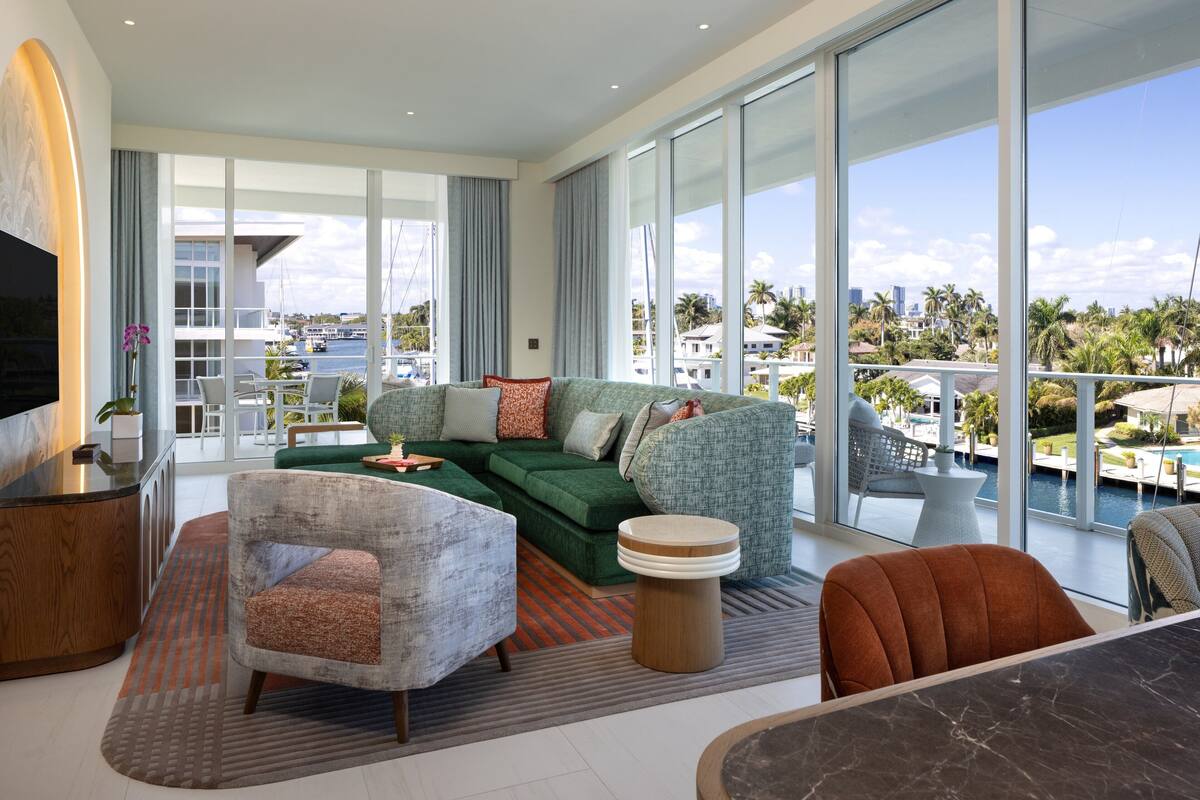 Amplia sala de estar con un sofá verde, una mesa de centro de madera y un televisor sobre un mueble. Amplias ventanas ofrecen vistas a la ciudad y un puerto deportivo.
