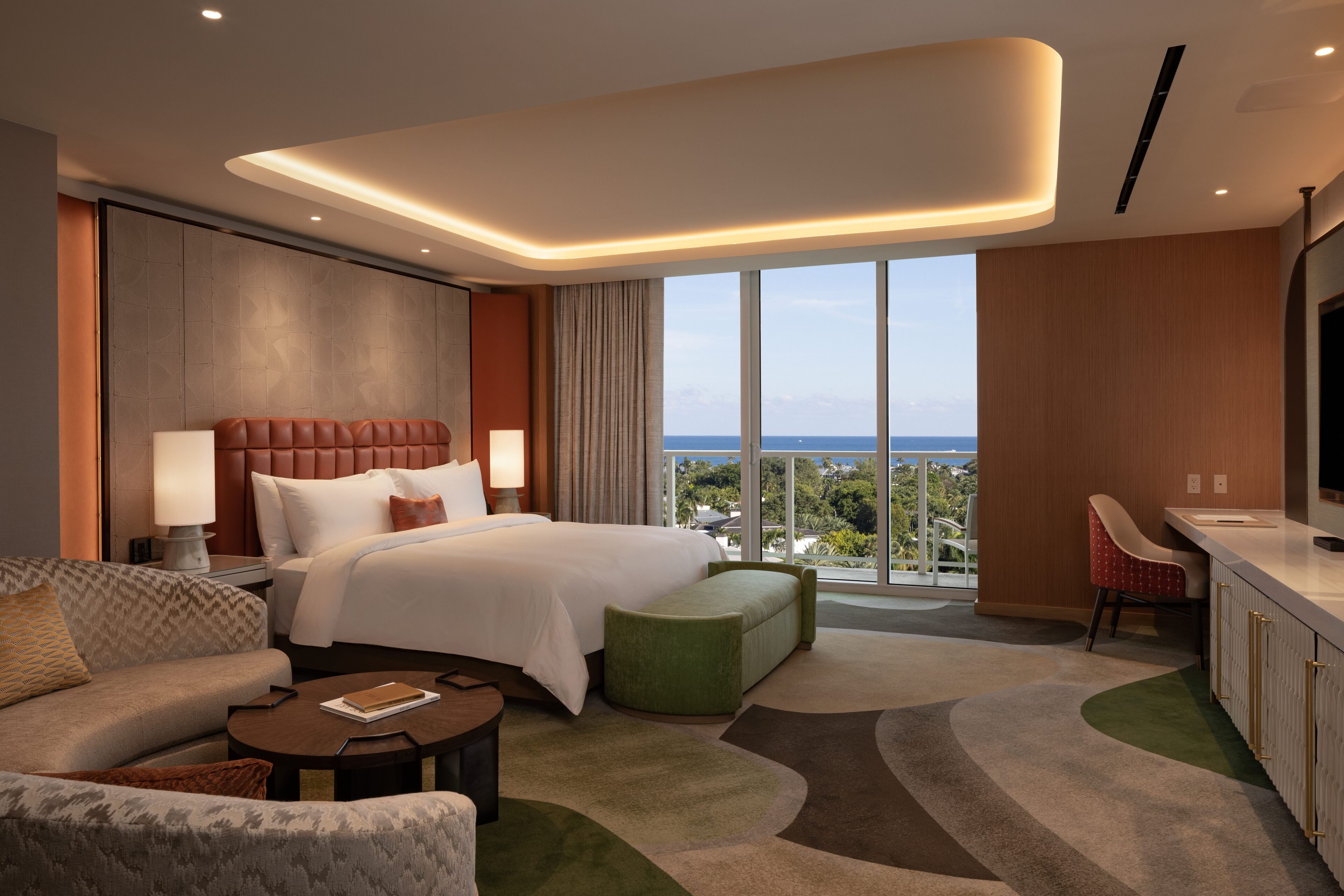 Una habitación de hotel moderna con una cama grande, una zona de estar con sofá y sillón, una mesita y vistas a la ciudad.