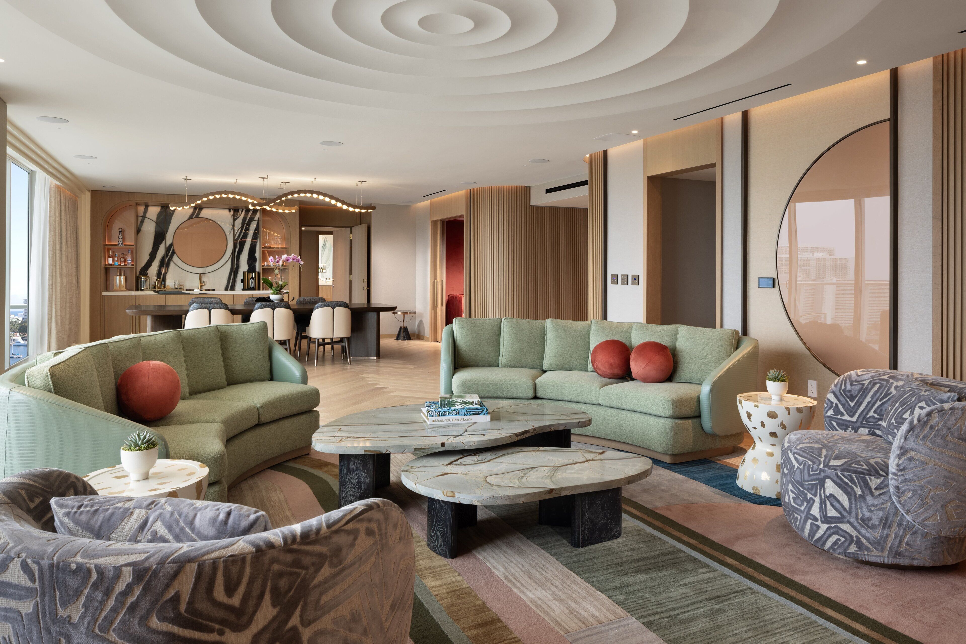 A modern living room with a green sofa, a patterned armchair, and a coffee table. There is a dining area in the background with a large mirror and a bar.