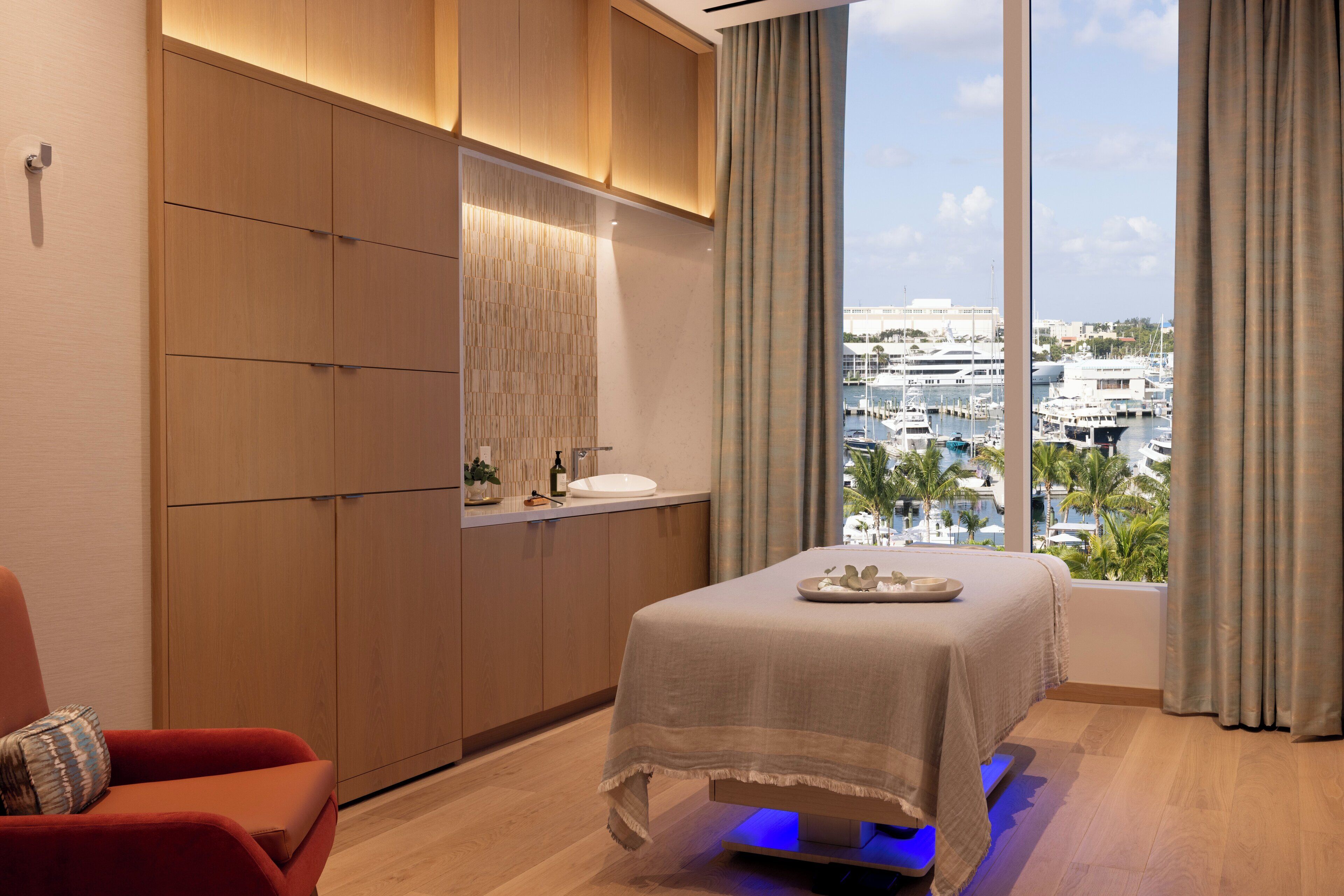 A modern bedroom with a large bed, a massage table, a red chair, and a view of a marina.