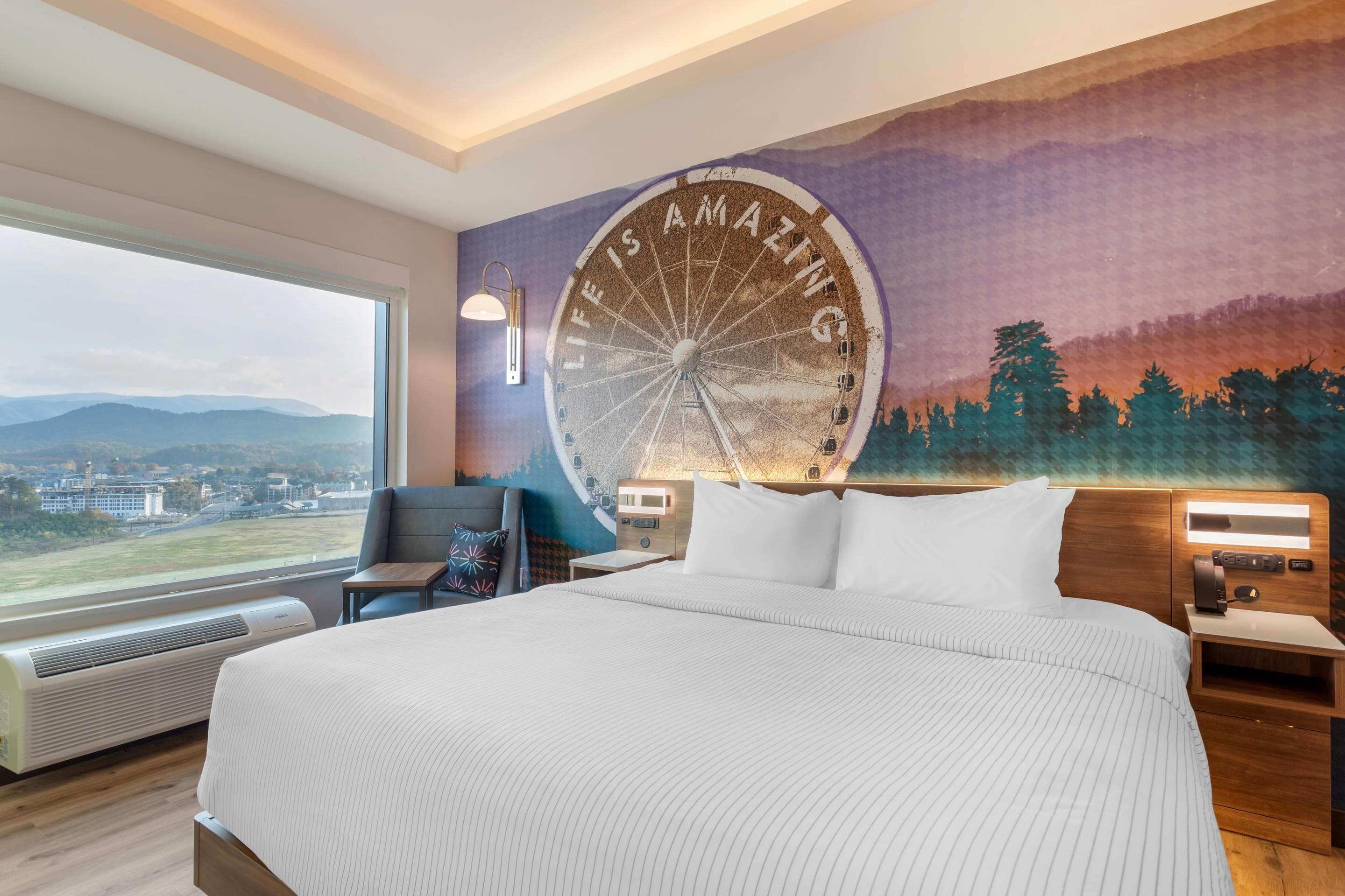 A hotel room with a large bed, a view of the city through a window, a wall mural with the text "LIFE IS AMAZING," and a wall-mounted clock.