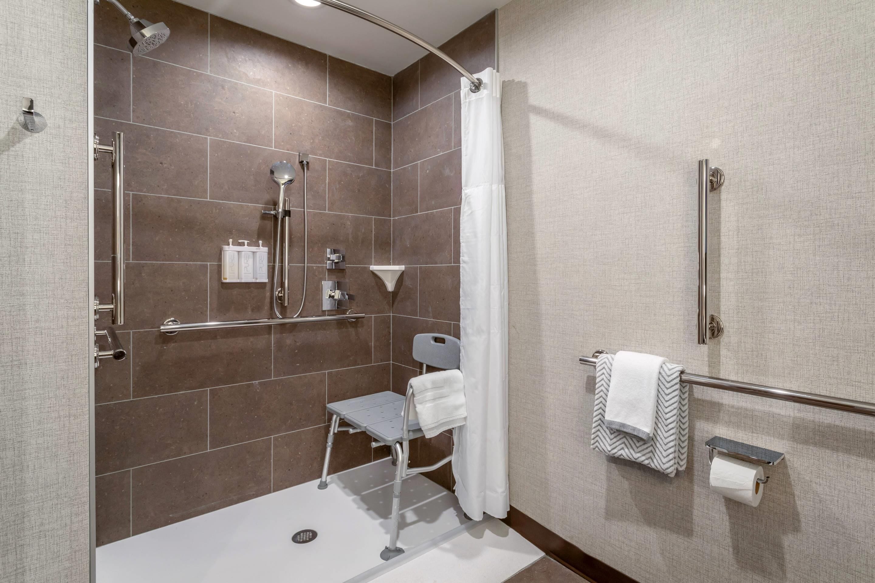 Un baño moderno con ducha, barras de apoyo, un taburete, toallero y soporte para papel higiénico.