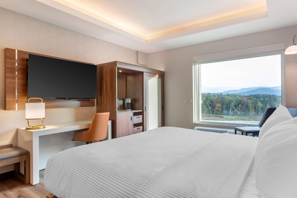 A modern hotel room with a large bed, a desk with a chair, a television, and a window with a mountain view.
