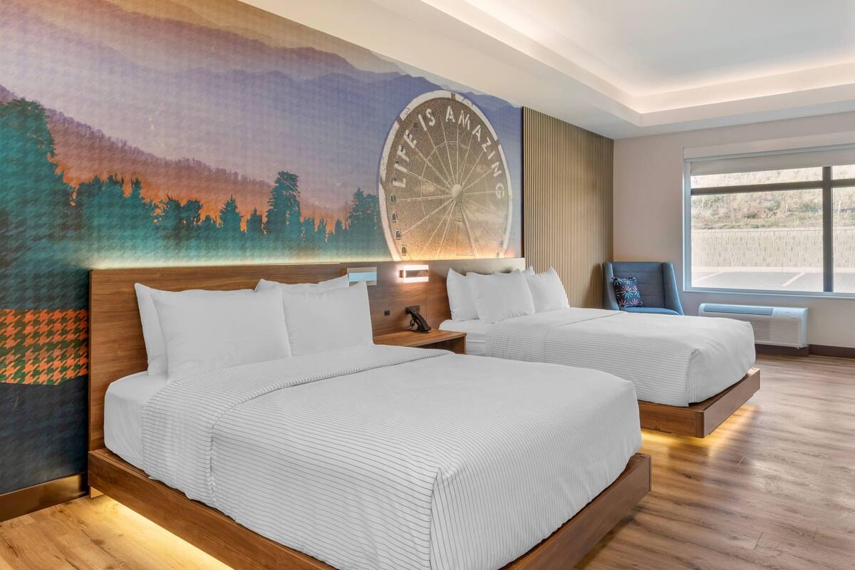 Ett hotellrum med två sängar, en väggmålning, ett stort fönster och utsikt över parkeringsplatsen.