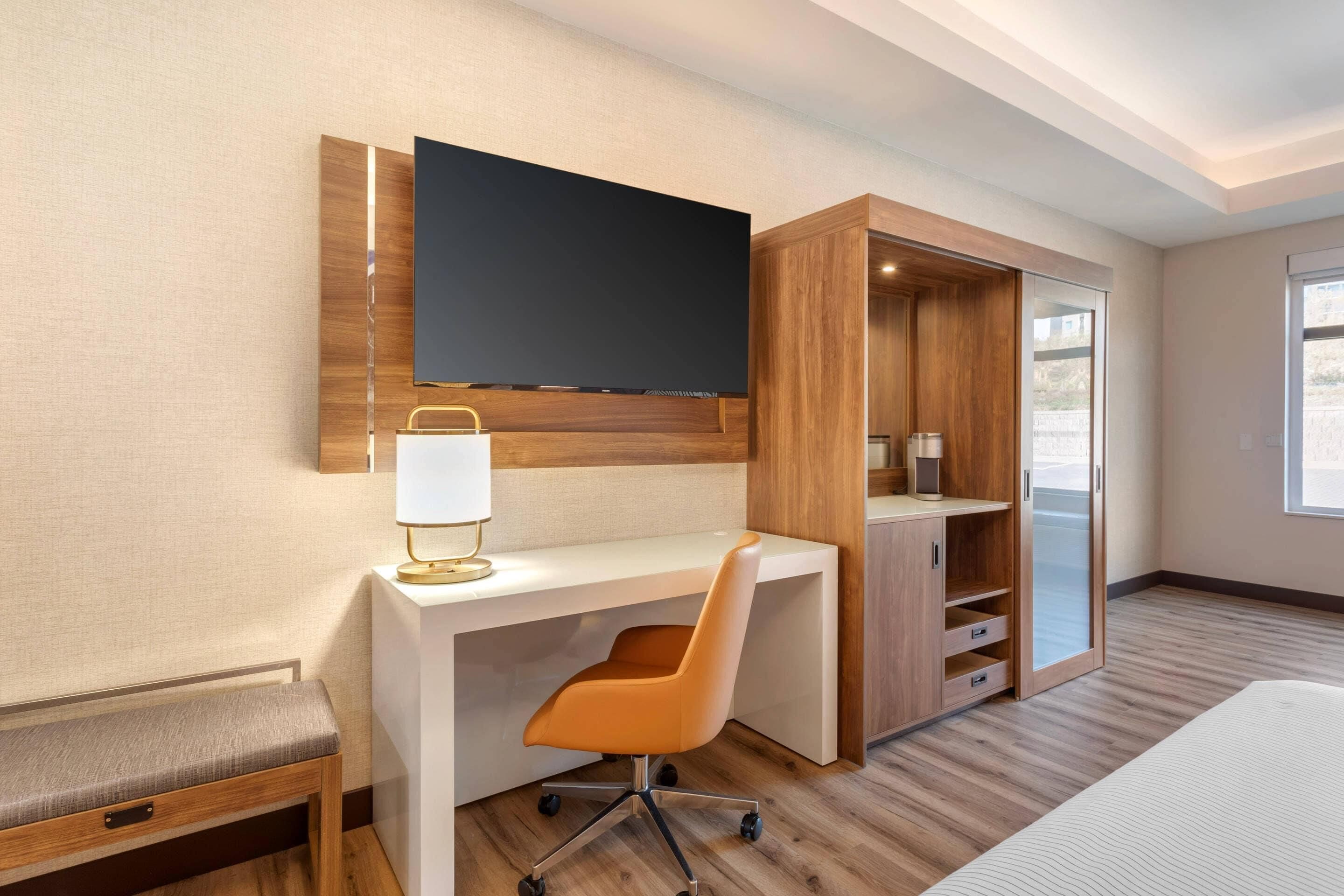 A hotel room with a wooden desk, a TV mounted on the wall, a chair, a bench, and a wardrobe.