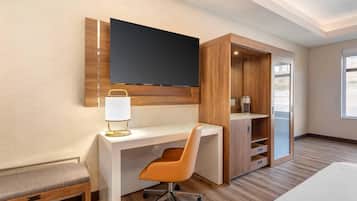 A hotel room with a wooden desk, a TV mounted on the wall, a chair, a bench, and a wardrobe.