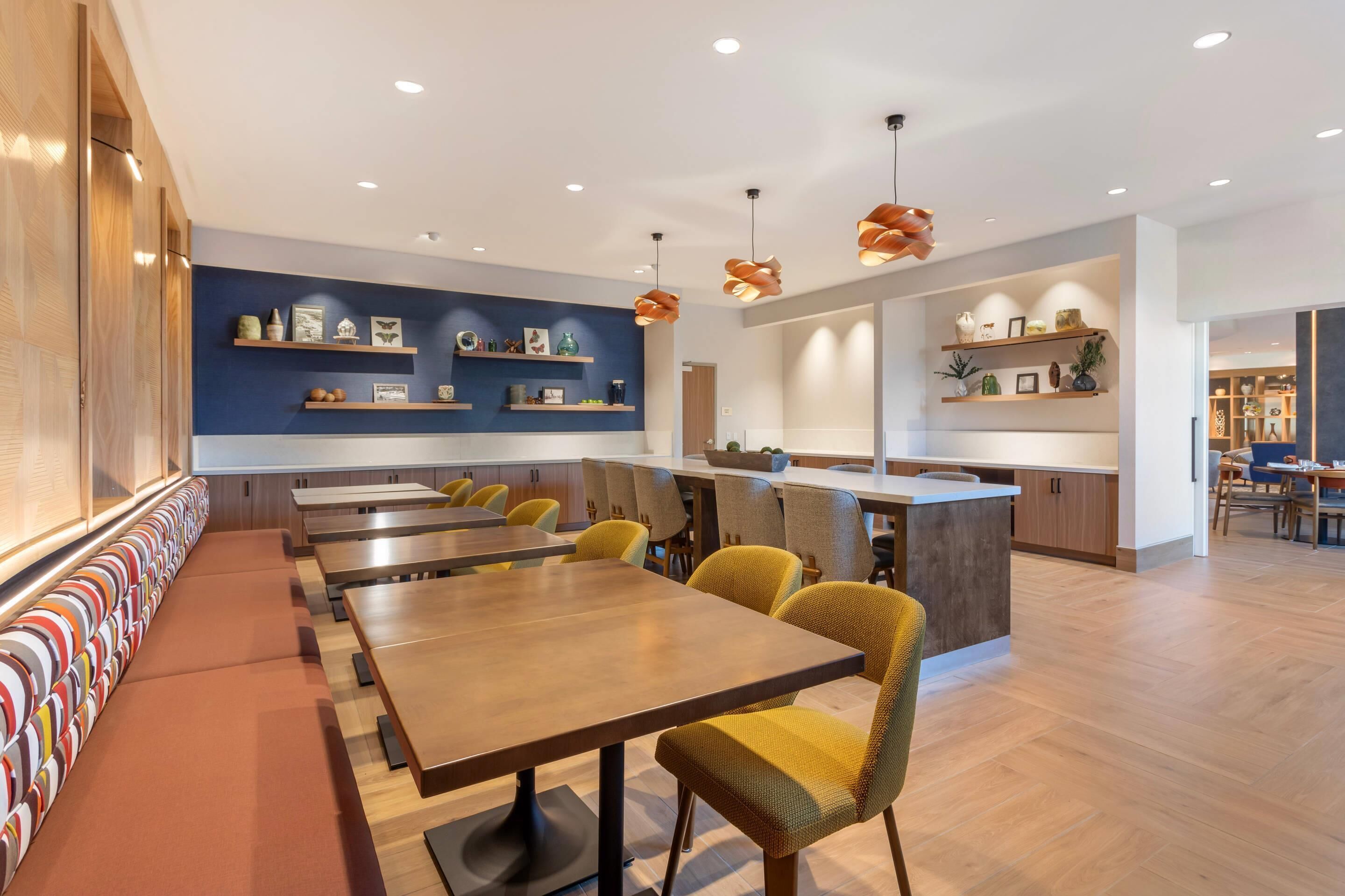 A modern dining area with wooden tables and yellow chairs, a long bench with colorful cushions, and shelves with decorative items on the walls.