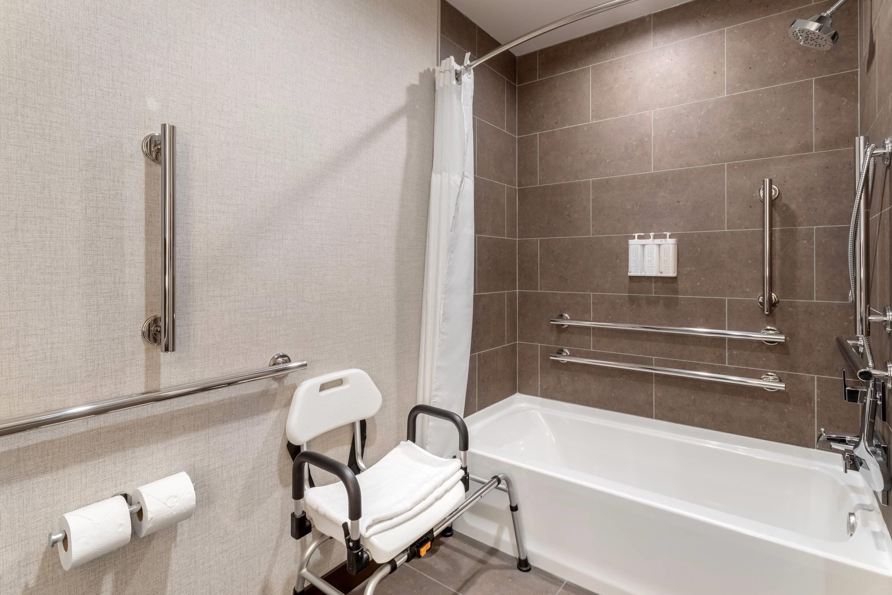A bathroom with a bathtub, a shower, a handrail, a white chair, and a toilet paper holder.