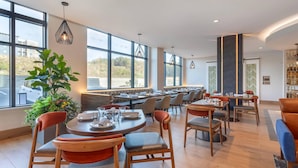 Un comedor moderno con mesas redondas, sillas acolchadas y grandes ventanales con vistas al exterior.