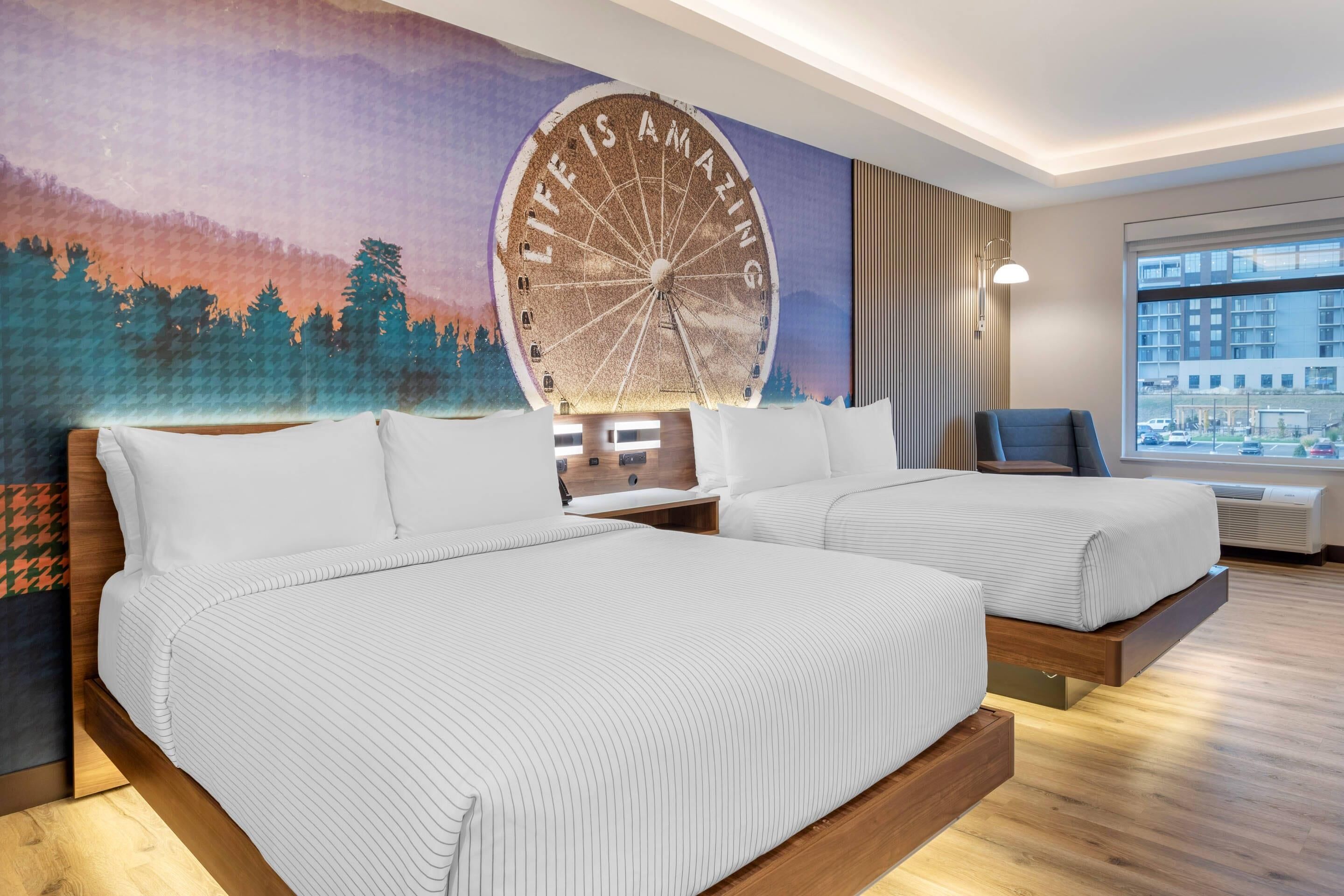 A hotel room with two beds, a mural with a Ferris wheel, and a city view through the window.