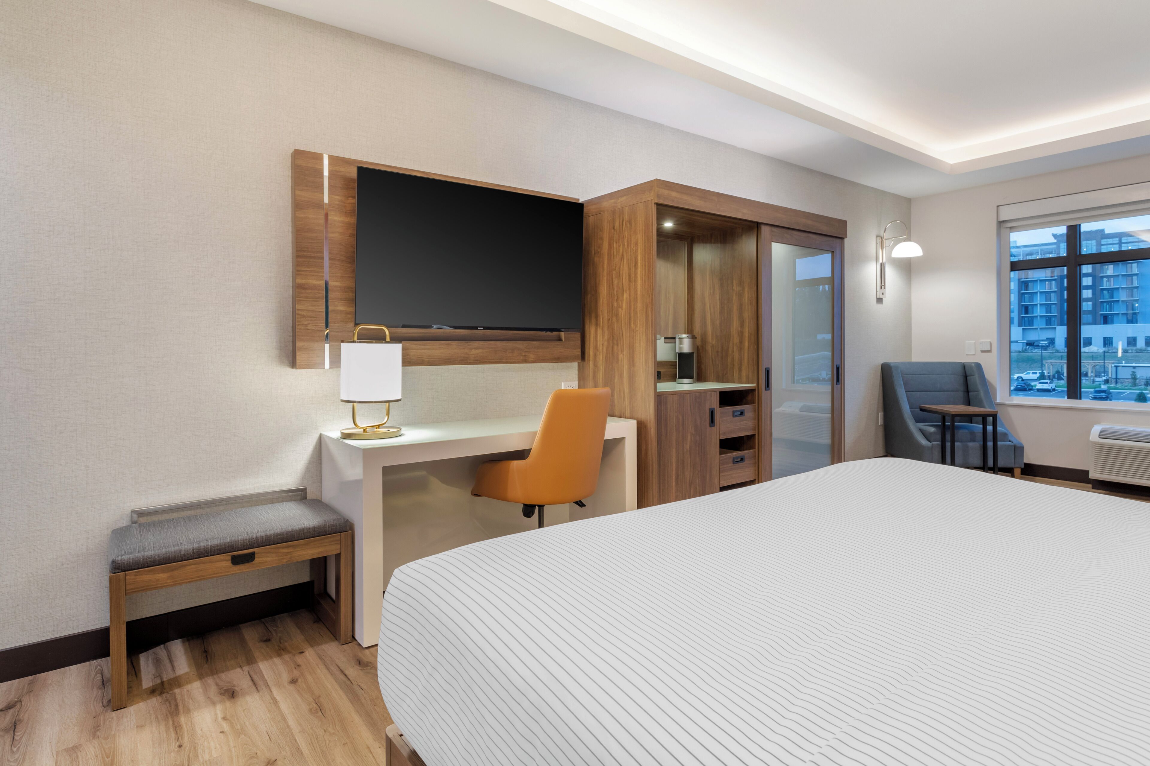 A hotel room with a large bed, a desk with a chair, a television mounted on the wall, and a window with a view of buildings.