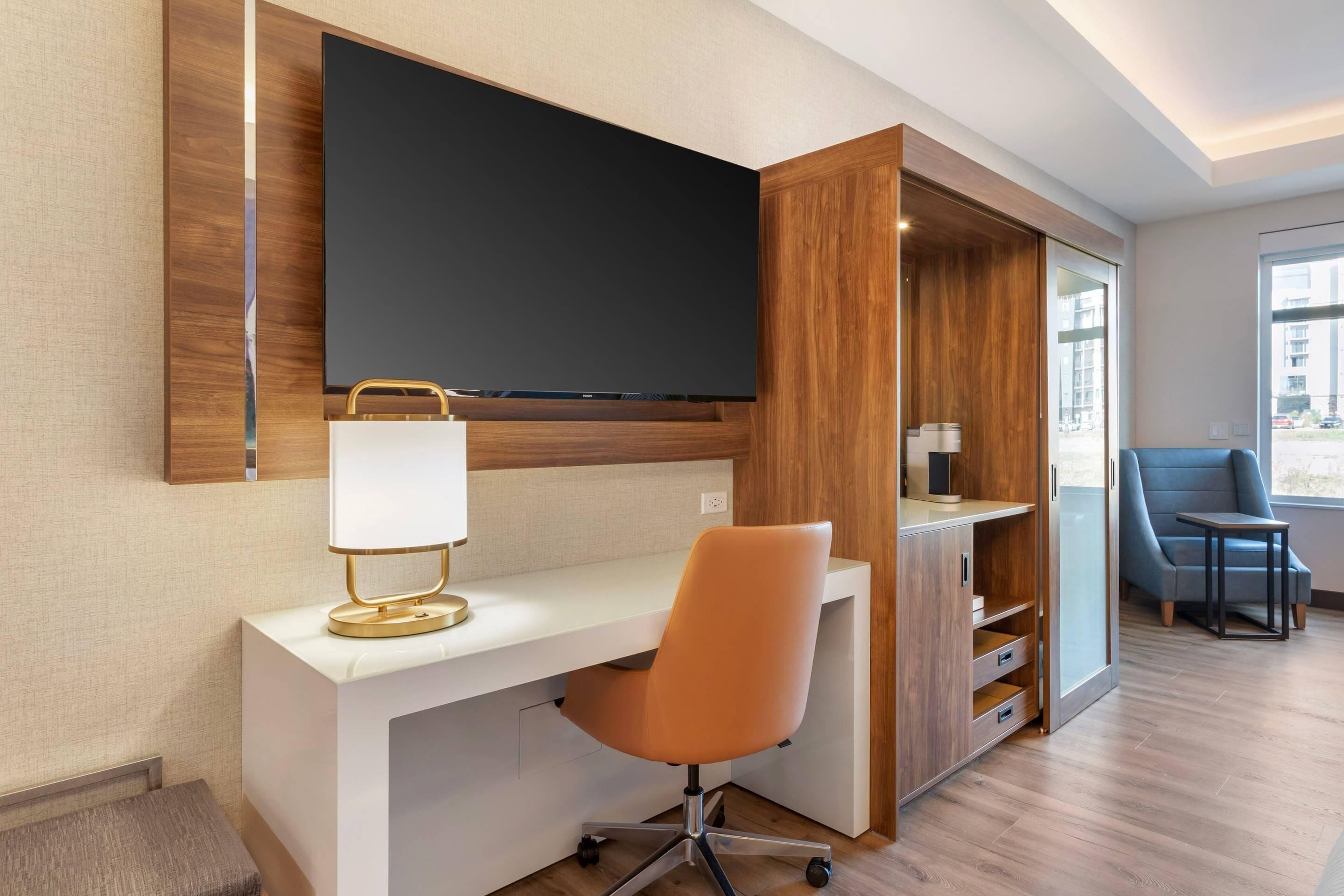A modern hotel room with a large flat-screen TV mounted on the wall, a white desk with a gold lamp, an orange office chair, and a blue armchair by a window.