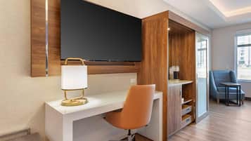 Ein modernes Hotelzimmer mit einem großen Flachbildfernseher an der Wand, einem weißen Schreibtisch mit goldener Lampe, einem orangefarbenen Bürostuhl und einem blauen Sessel am Fenster.