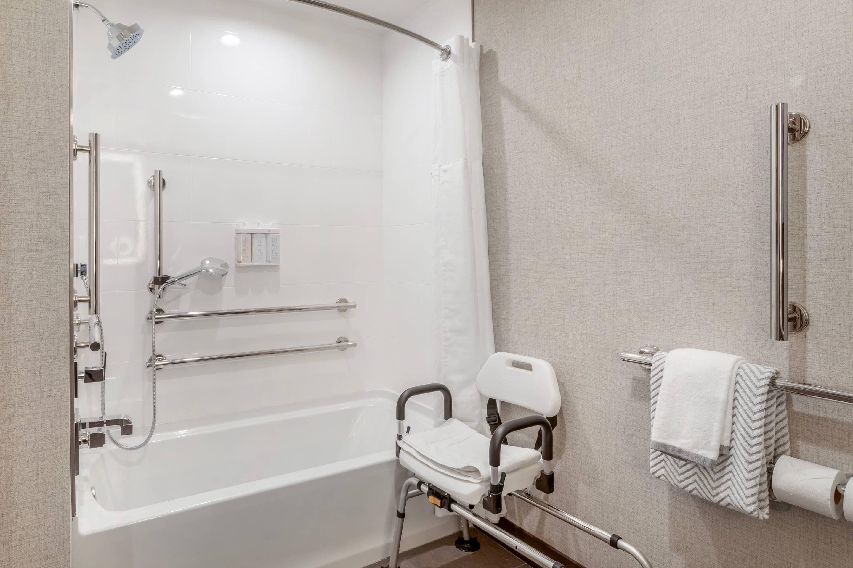 A bathroom with a white bathtub, a shower with a handheld showerhead, a grab bar, a white chair, and a towel rack with a towel.