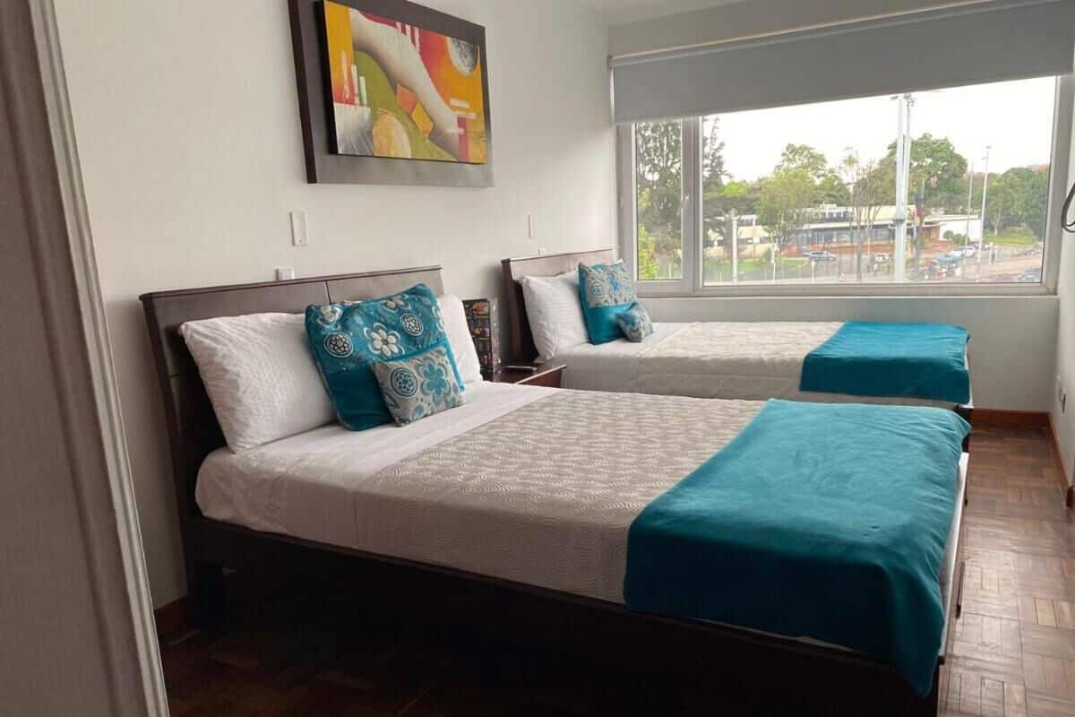 A hotel room with two beds, each with a white bedspread and teal accents. There is a window providing a view of the outside.