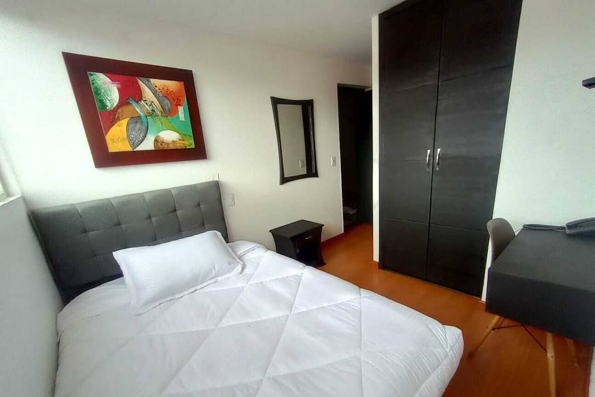 A hotel room with a bed, a desk, a wardrobe, and a painting on the wall.