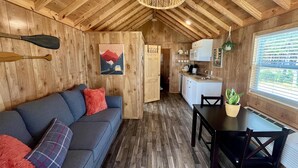A cozy cabin interior with a wooden ceiling, a sofa with red and plaid cushions, a dining table with chairs, a kitchen area, and a window with blinds.