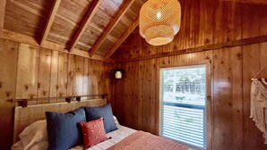 A wooden cabin bedroom with a bed, pillows, a hanging light fixture, and a window with blinds.