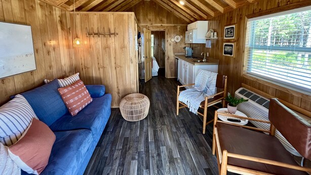 A cozy cabin interior with a wooden ceiling, a blue sofa, a wicker chair, a small kitchenette, and a window with blinds.