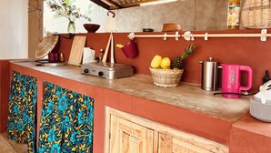 A kitchen with a gas stove, a sink, a hanging basket with fruits, a pink kettle, and a metal container.