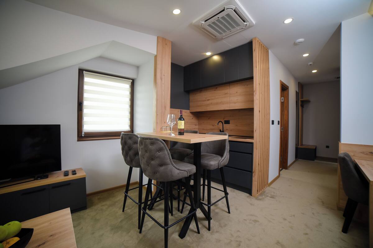 A modern kitchen with a dining area, featuring a wooden table and bar stools, a TV, and a large window.
