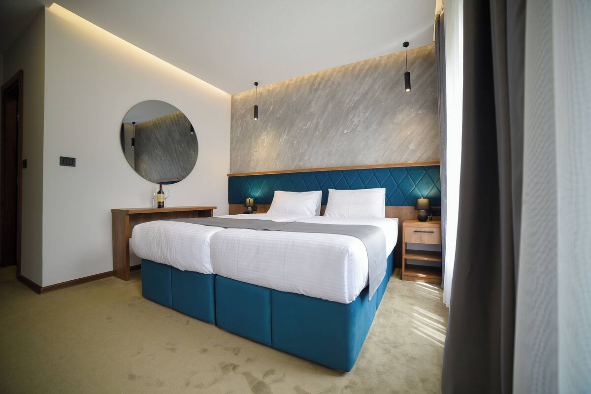 A modern hotel room with a large bed, a round mirror, a wooden headboard, and a nightstand with a lamp.