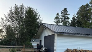 An outdoor patio with a table and chairs, a barbecue grill, and a stack of firewood against a building with a garage. - Large newly renovated house.  Cozy and convenient.  (Valemount)