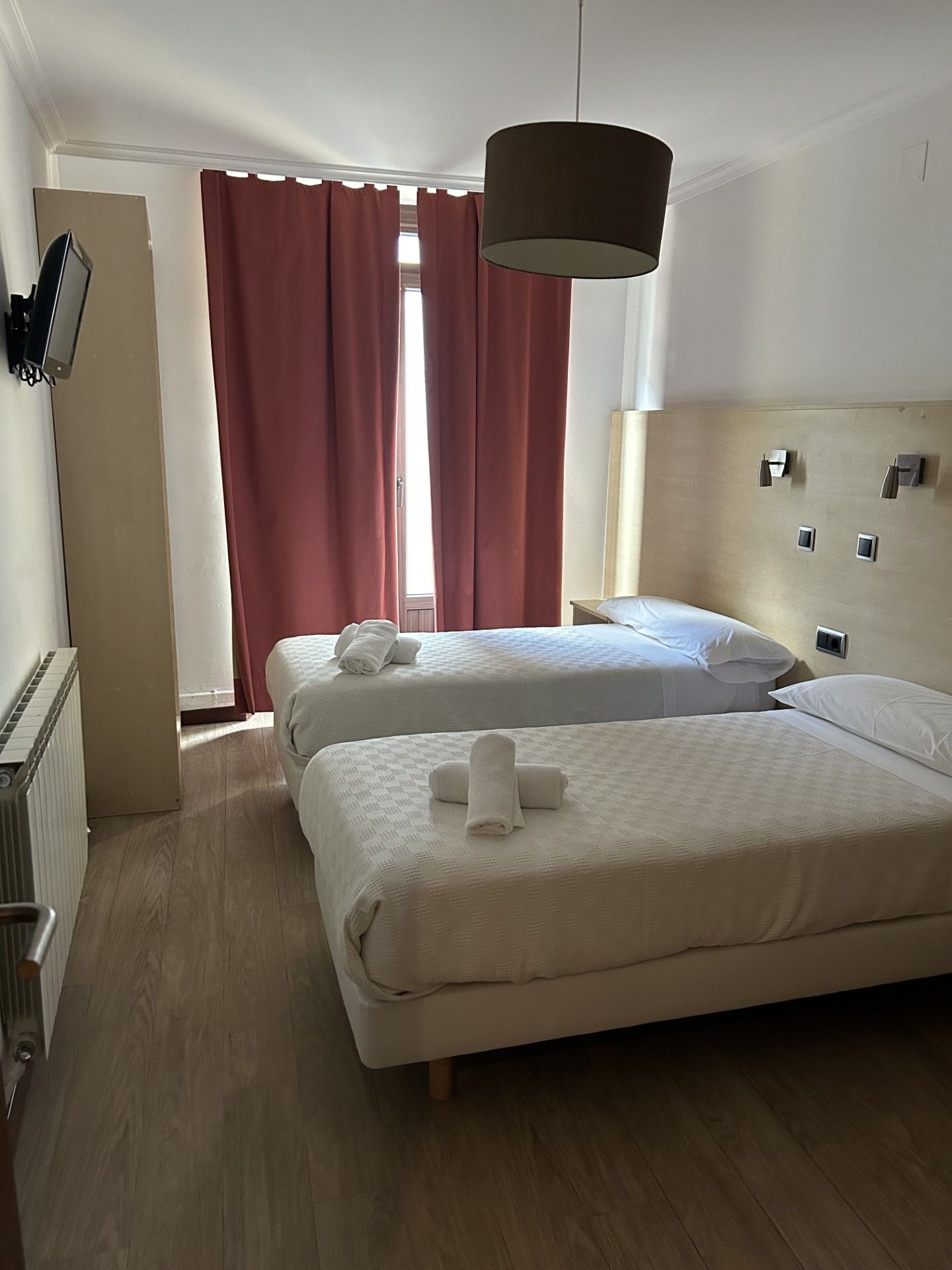 Quarto de hotel com duas camas, uma janela grande com cortinas vermelhas, uma luminária no teto e uma TV fixada na parede.