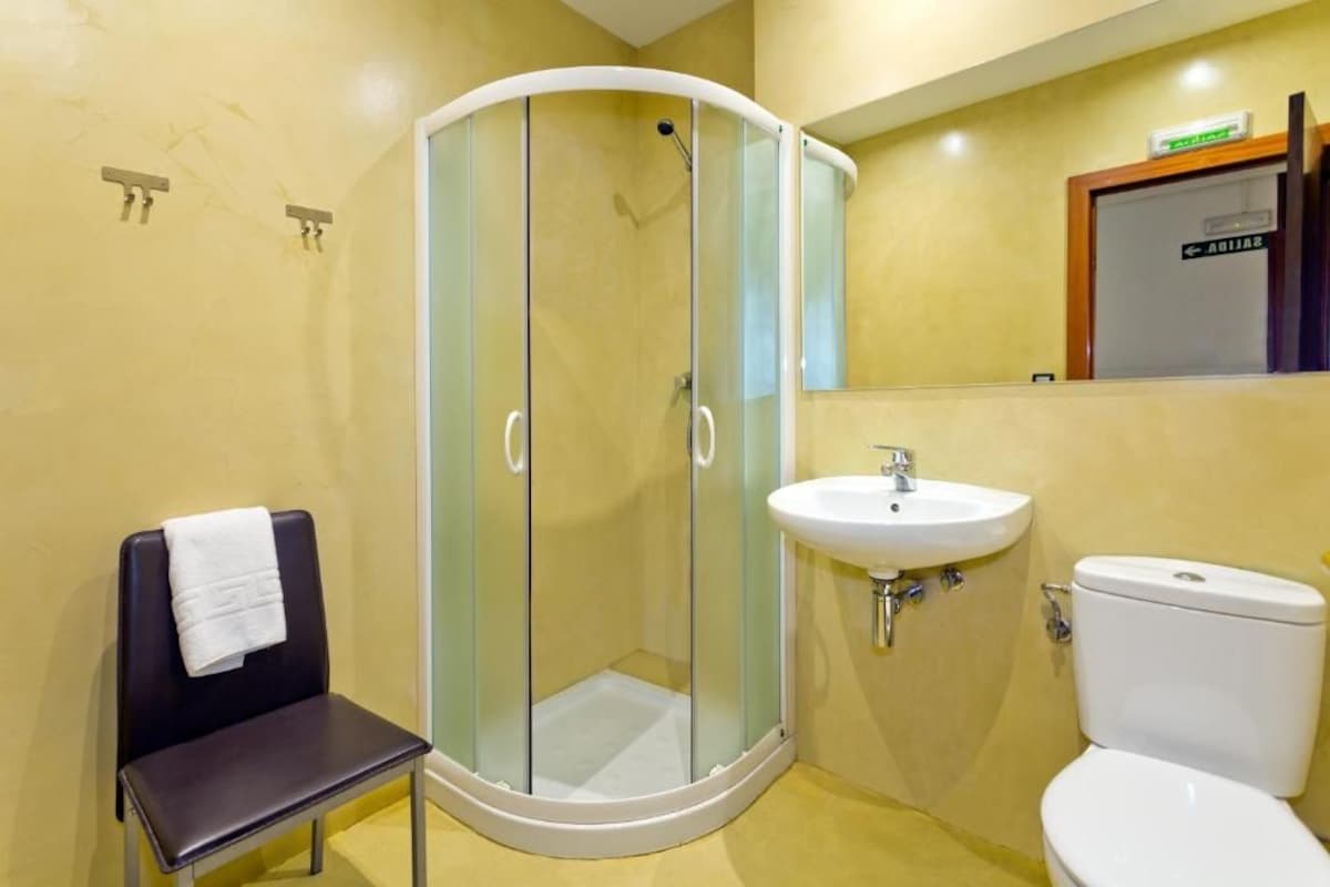 A bathroom with a glass-enclosed shower, a sink, a toilet, and a chair with a towel.