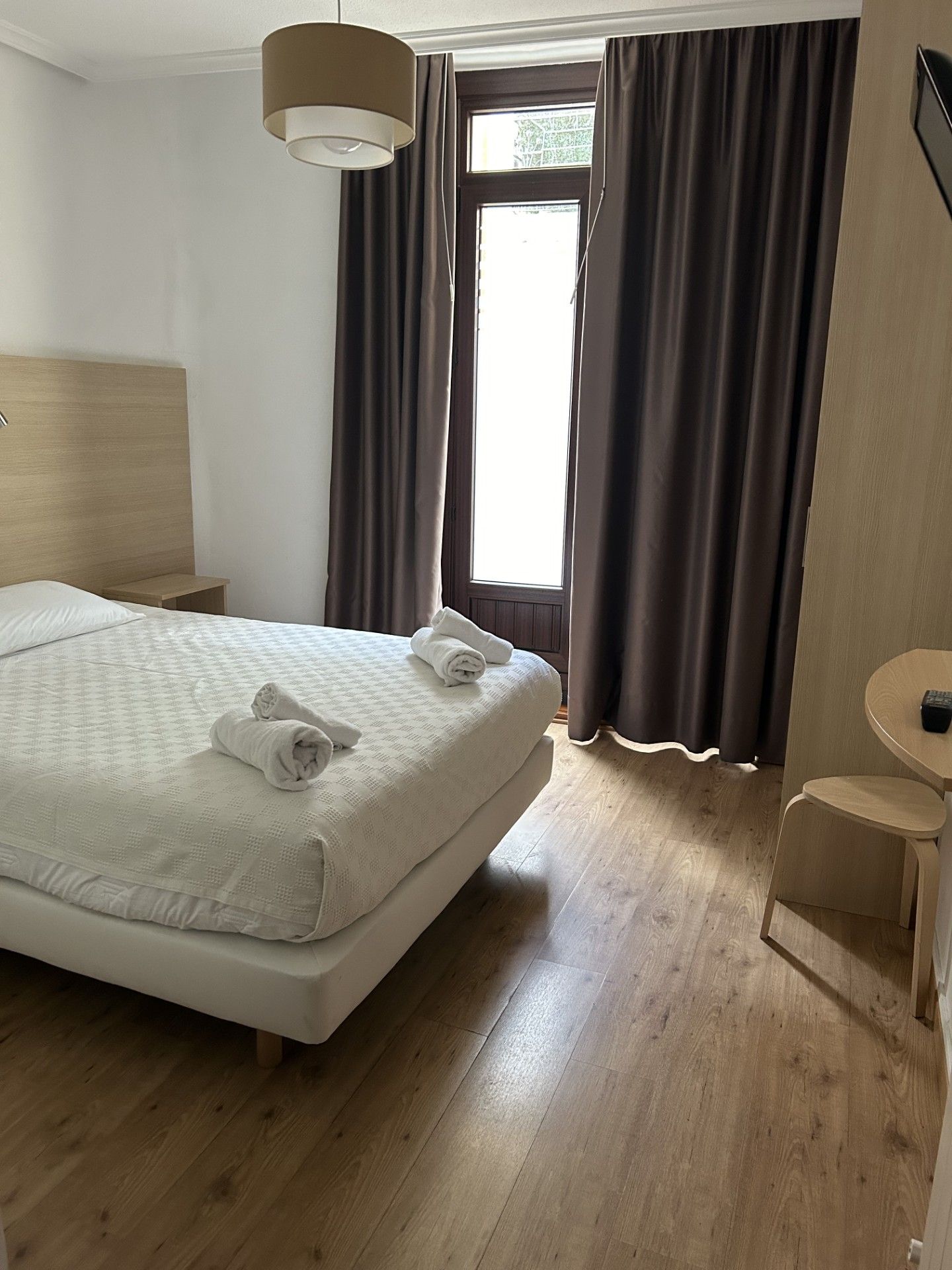 Ein ordentlich eingerichtetes Hotelzimmer mit einem großen Bett, einem Nachttisch, einem kleinen runden Tisch und einem Fenster mit Vorhängen.