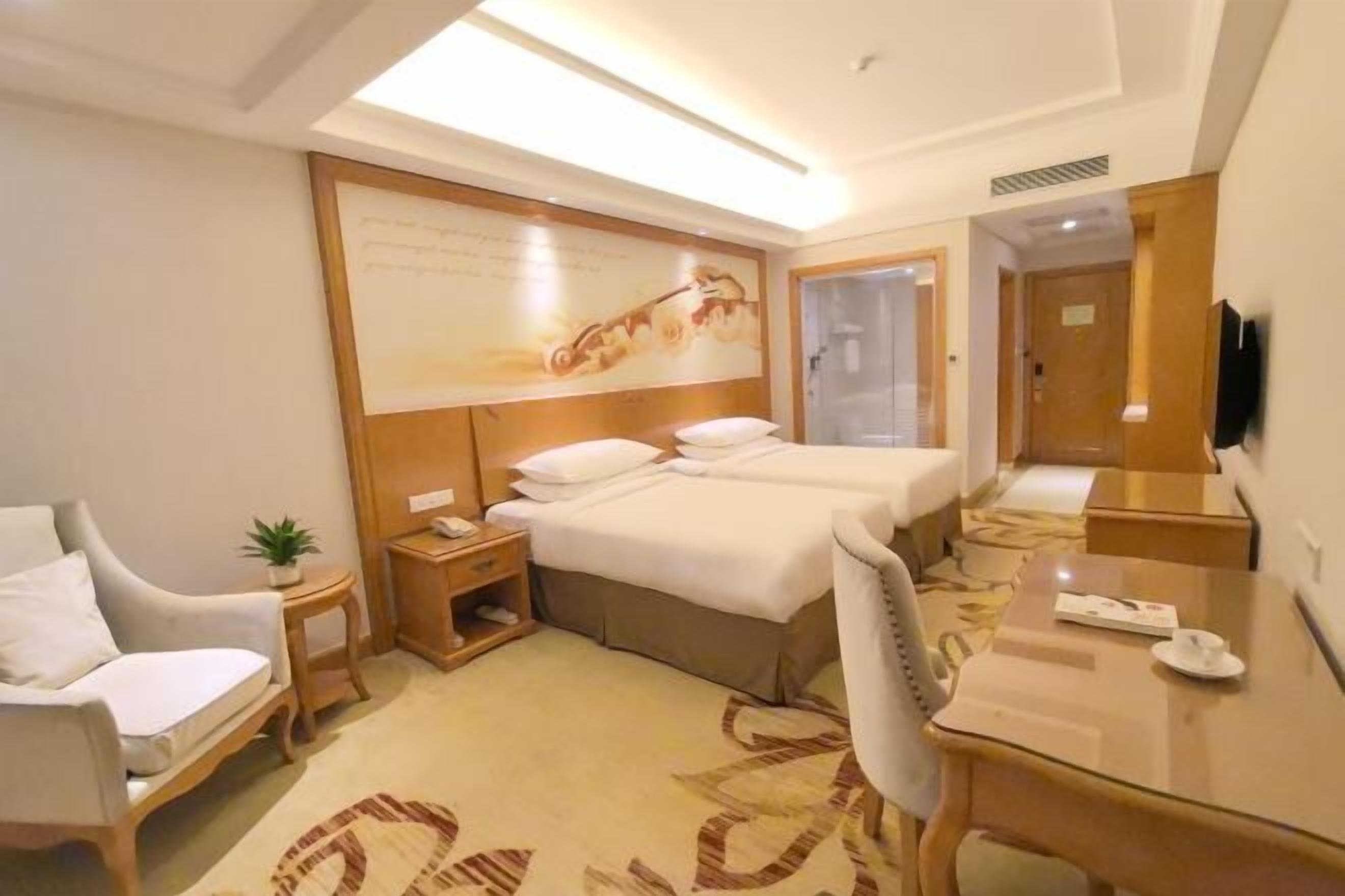 A hotel room with a bed, a desk, a chair, a nightstand, a small table with a plant, and a wall-mounted TV.