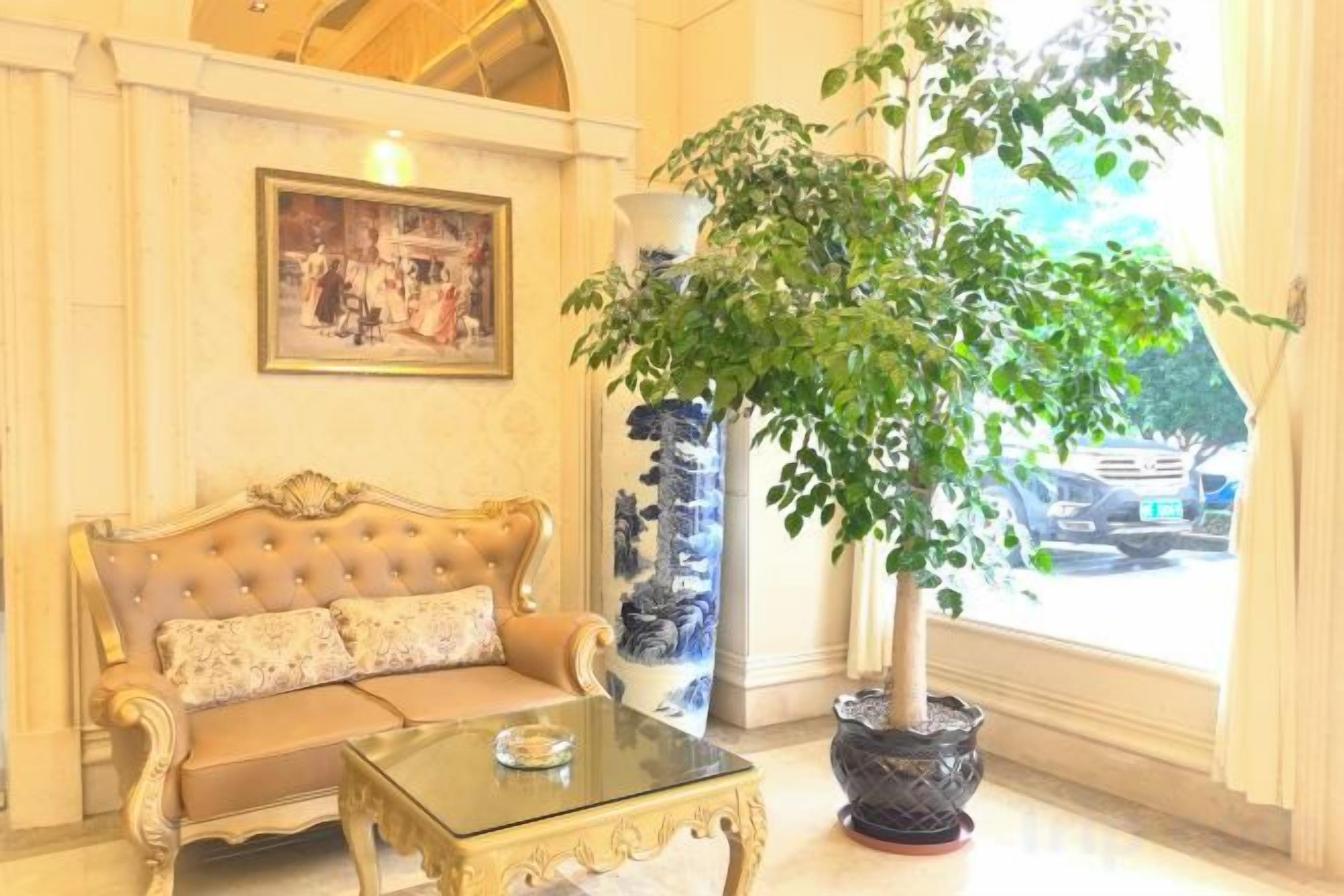 A room with a sofa, a glass coffee table, a large potted plant, a vase, and a painting on the wall.