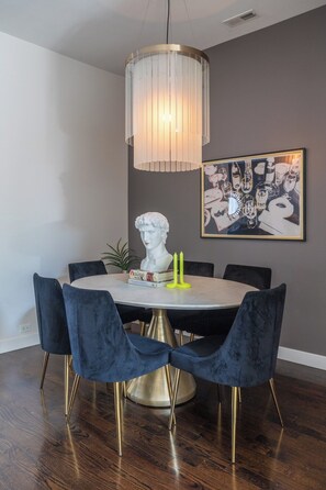 A dining area with a round table, dark blue velvet chairs, a gold base, a modern pendant light, and a bust sculpture.