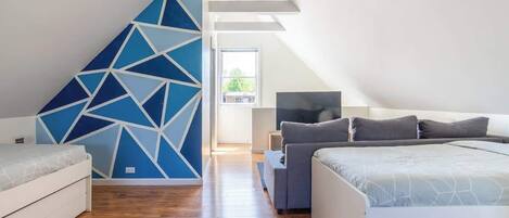 A modern bedroom with a geometric-patterned wall, a bed with a geometric-patterned bedspread, a grey sofa, and a TV on a side table.