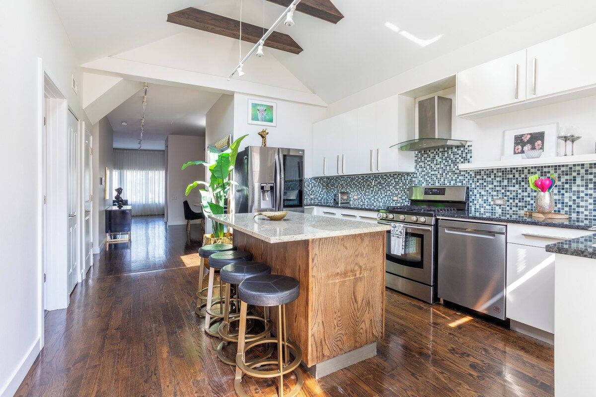 A modern kitchen with a central island, bar stools, stainless steel appliances, and a tiled backsplash.