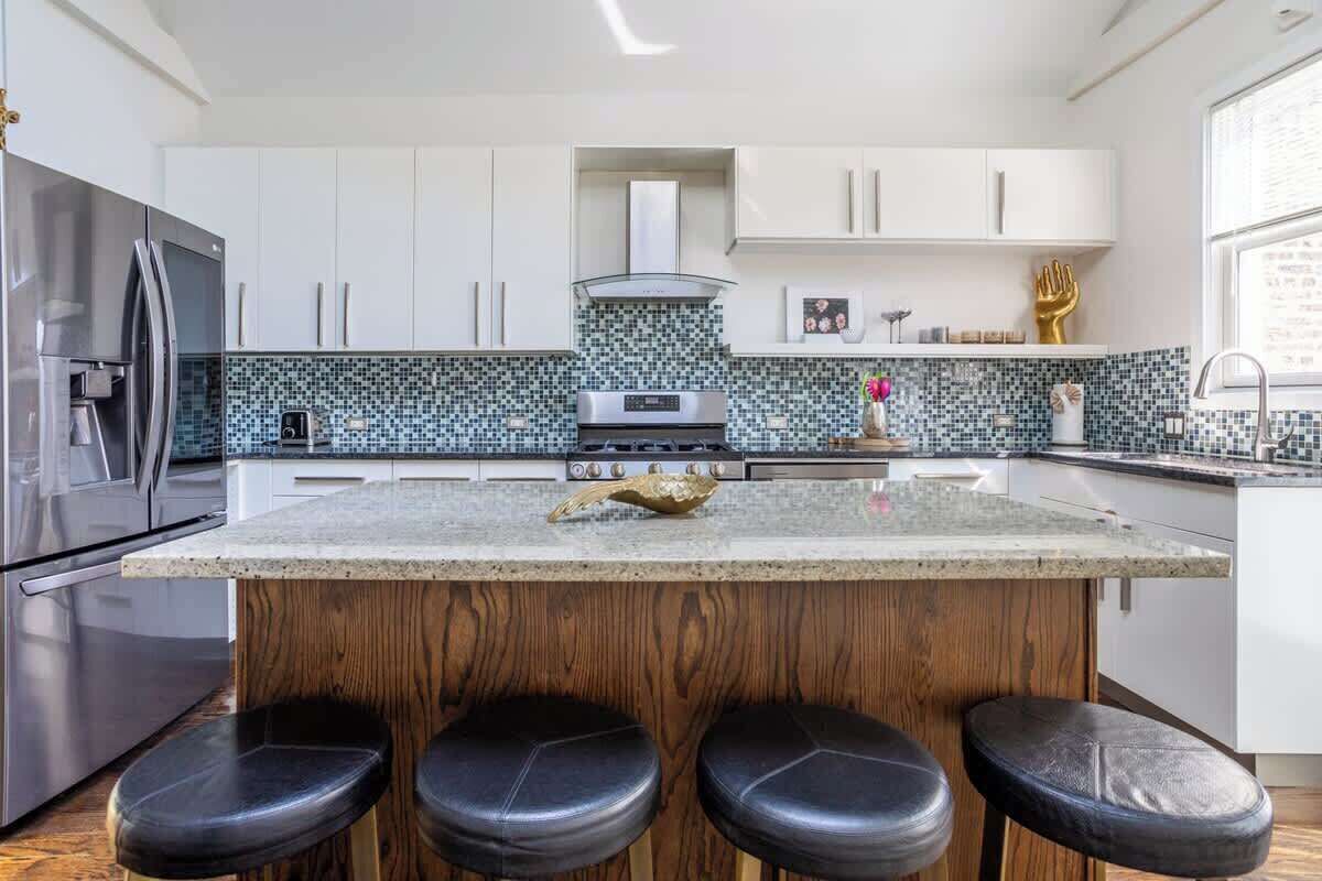 Una cocina moderna con armarios blancos, una isla central con taburetes, un refrigerador de acero inoxidable y un salpicadero con diseño.