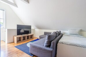 A modern living room with a flat-screen TV on a wooden stand, a grey sofa, and a bed with white bedding.