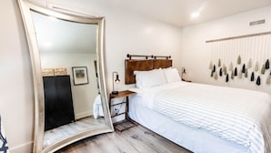 Un dormitorio con cama, un espejo grande, cabecera de madera, mesita de noche y un arreglo de plantas en la pared.