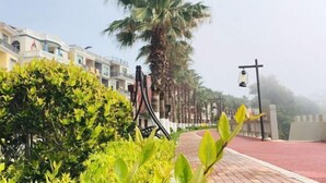 Un camino bordeado de palmeras y pavimento rojo, que conduce a una hilera de edificios de varios pisos con balcones.