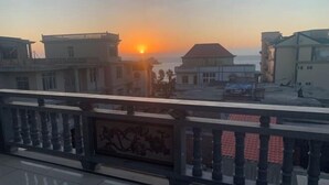 Un balcón con vistas a la puesta de sol sobre el mar, edificios al fondo y un suelo de baldosas.